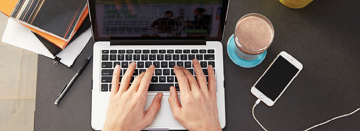 Person Typing On A Laptop With A Shake And Phone Next To Them